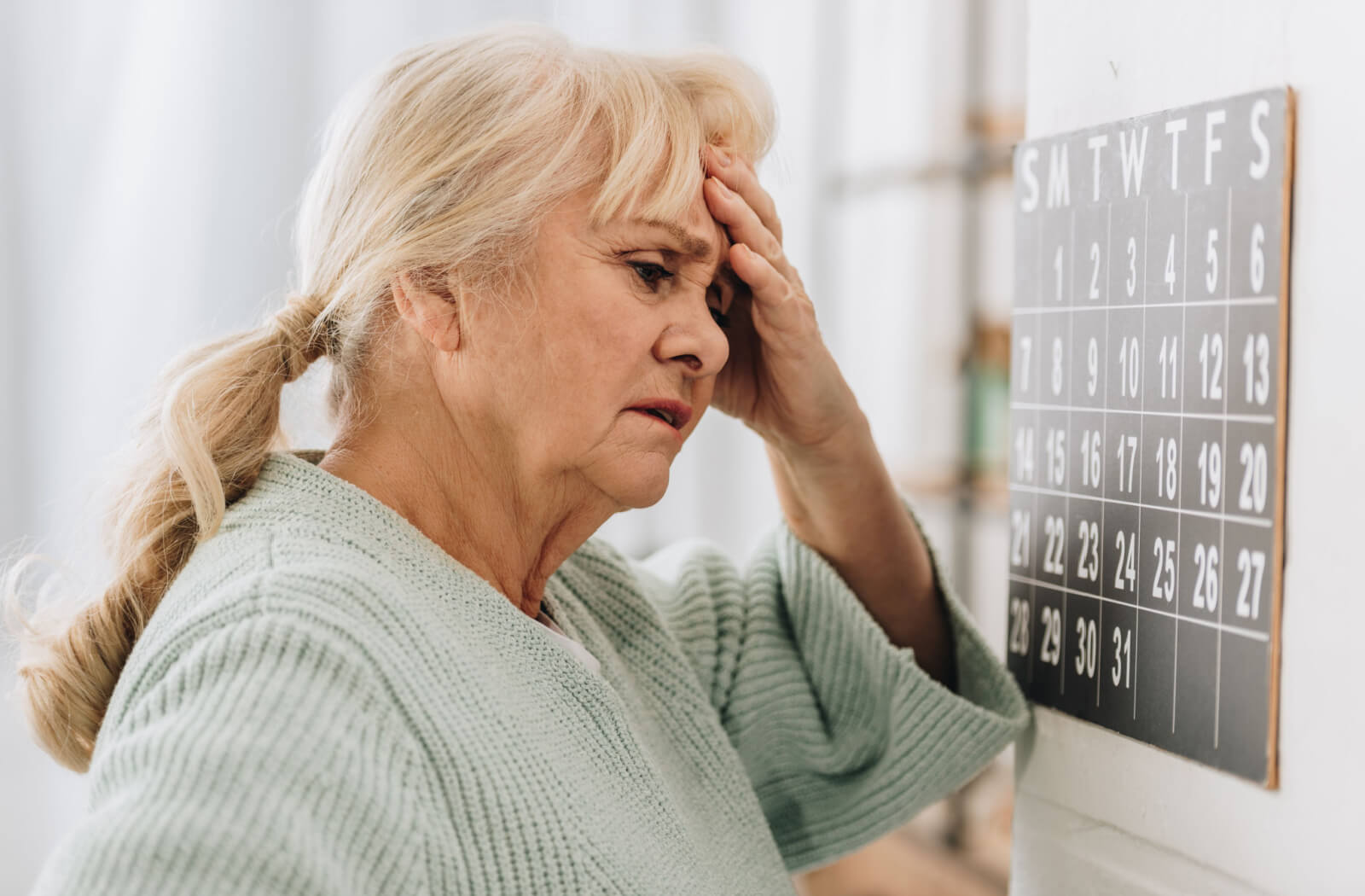 Standing before the calendar on the wall, the older woman furrows her brow, touching her head as she tries to recall the date