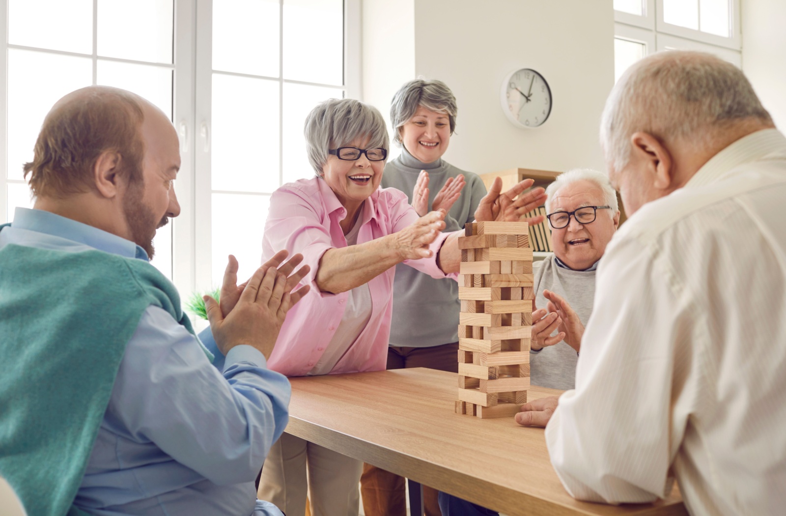 A group of older adults in personal care standing around a tower of wooden blocks during a fun game of Jenga.