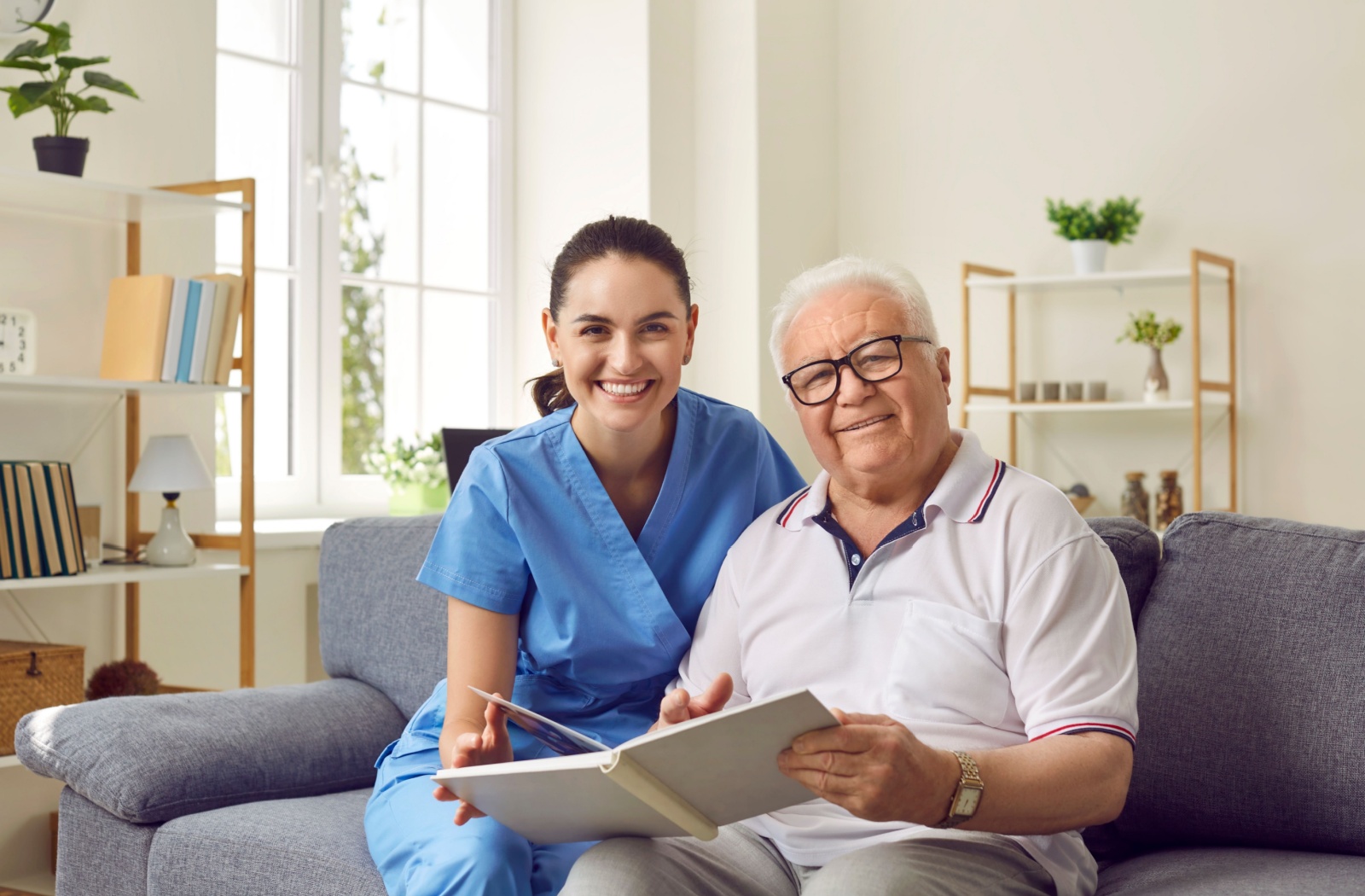 A caregiver and an older adult with dementia smiling while looking through a photo album on a couch in memory care.