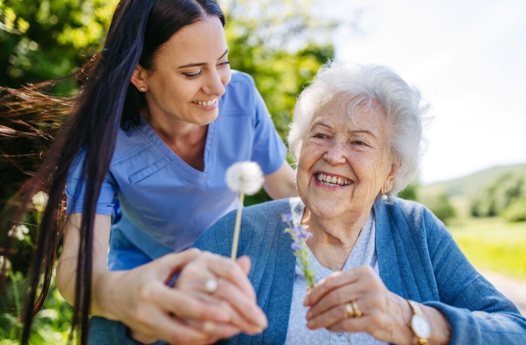 A caregiver and an older adult with dementia in memory care walking outdoors and smiling while holding a dandelion.