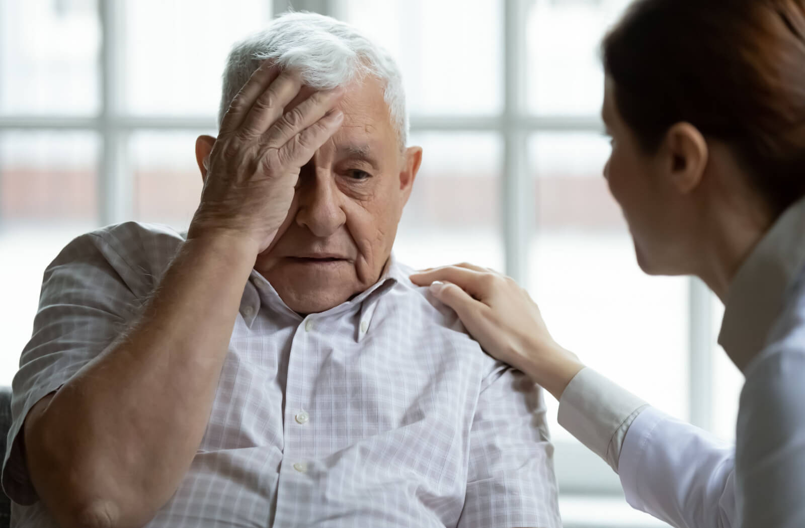 An older adult rubbing their forehead in frustration due to dementia while a caregiver reassures them.