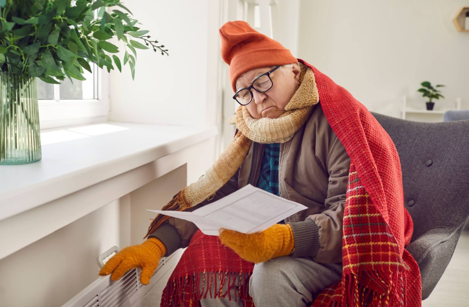 An older adult sitting near a heating vent bundled in warm clothes, a blanket, hat, and gloves while looking at a paper.