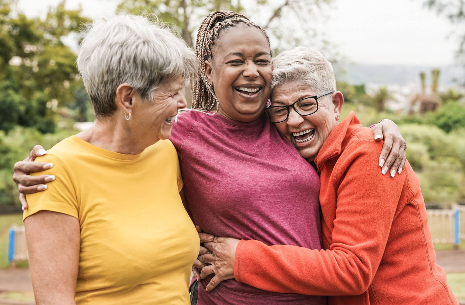 Three older friends hugging while on a walk through the park during the summer.