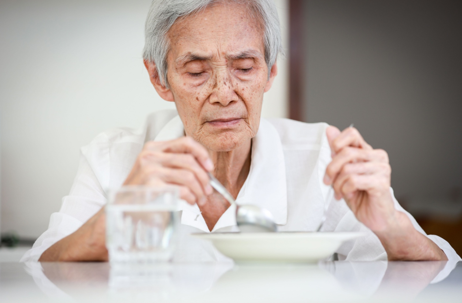 An uninterested older adult sits with a glass of water and a bowl of soup, stirring constantly due to a lack of appetite