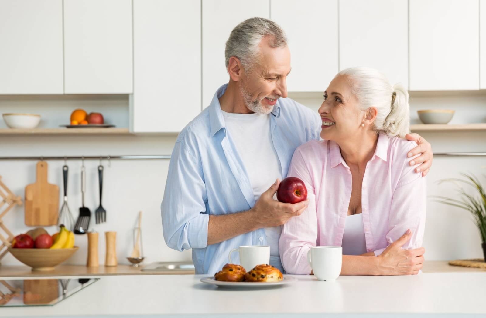 An older adult hands their smiling spouse an apple in the kitchen to encourage them to eat
