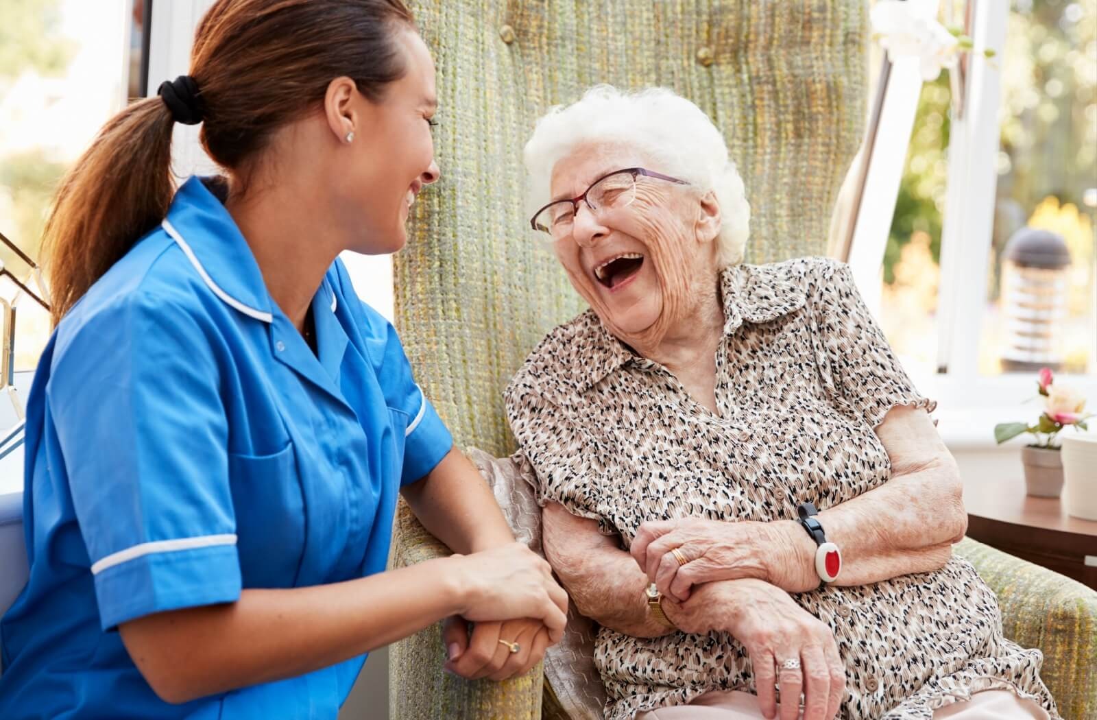 A caregiver smiles at a laughing memory care resident sitting in an armchair.
